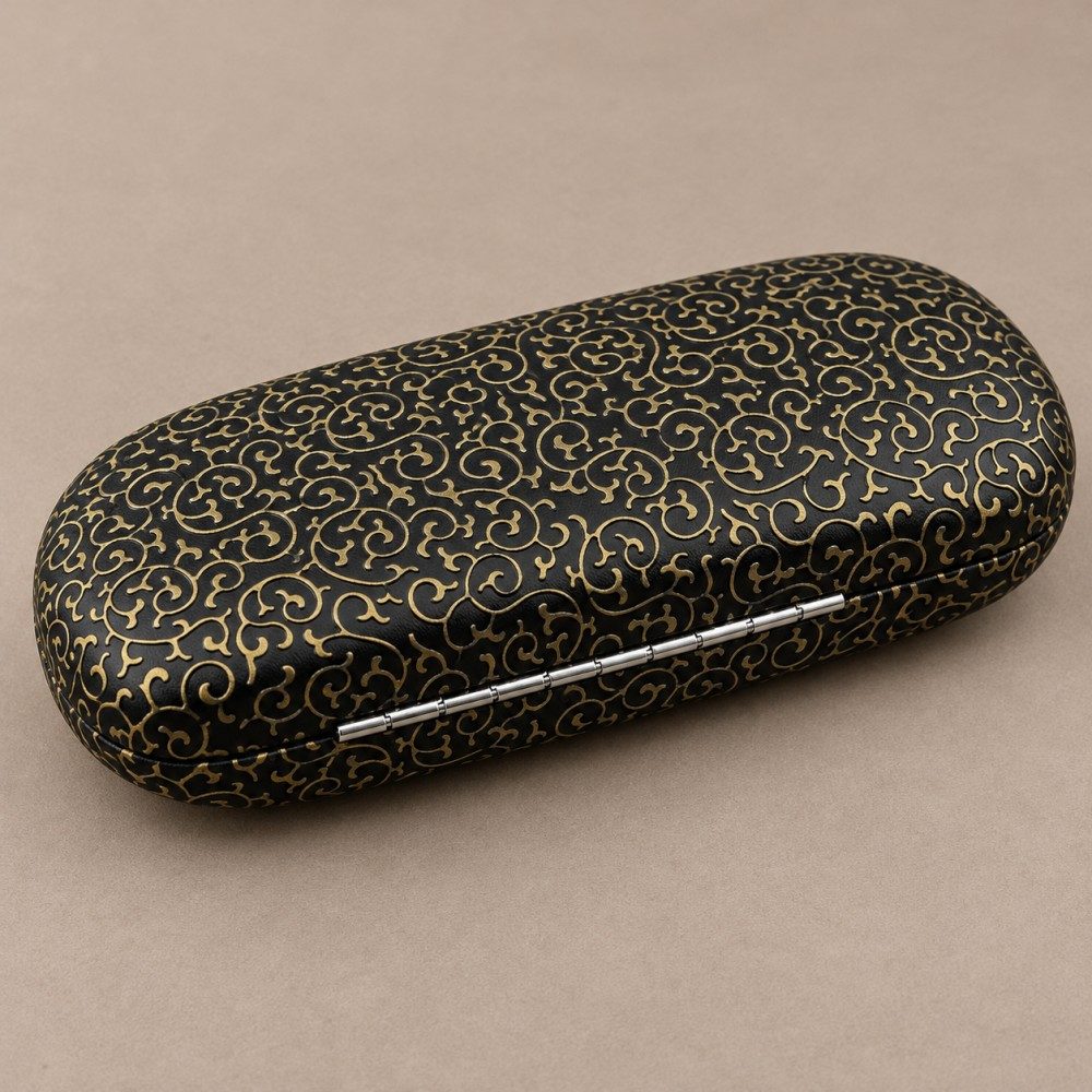 Black PU-covered metal eyeglass case with gold scroll detailing and a visible silver hinge on a taupe background.