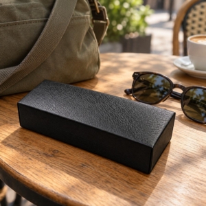 Black rectangular sunglasses case on a wooden cafe table beside sunglasses and an olive canvas bag.