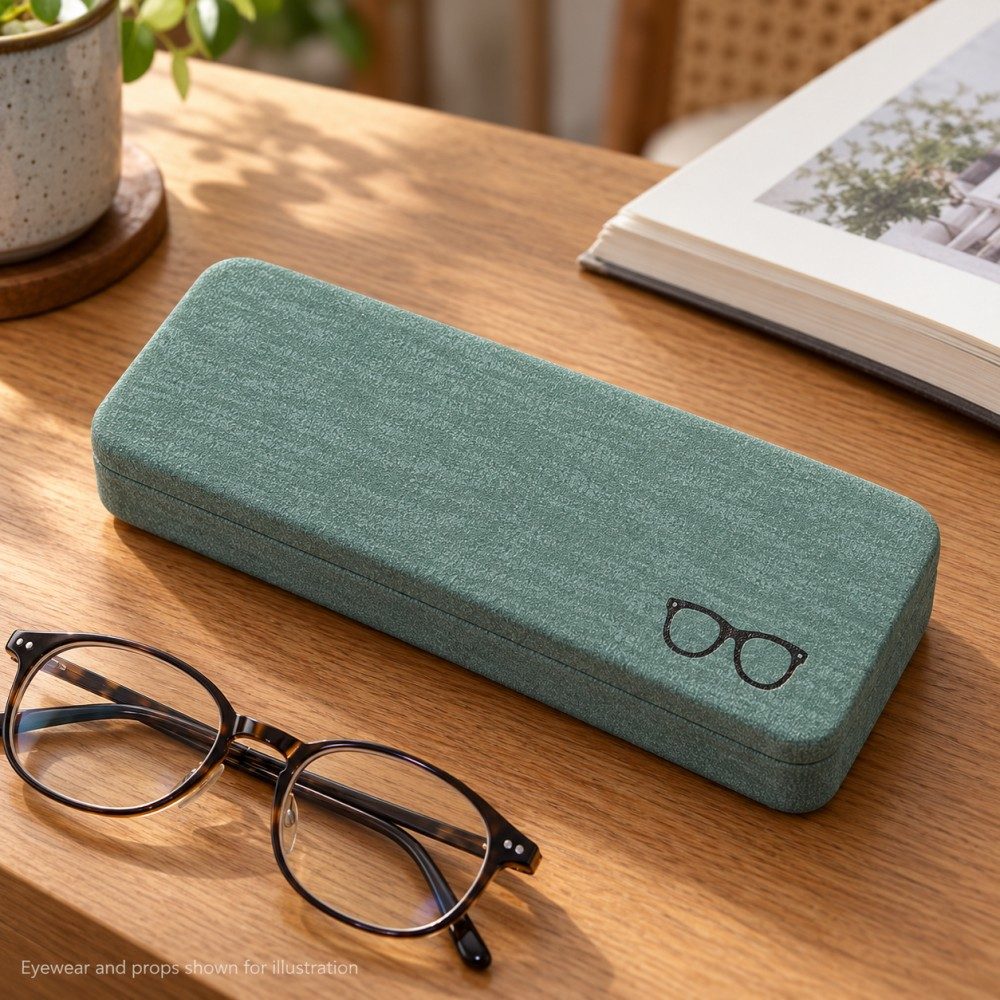 Pale green glasses case beside folded eyeglasses and an open book on a sunlit wooden reading desk.
