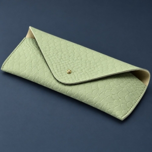 Light green scale-embossed sunglasses case with an envelope flap and small gold-tone stud on a dark blue-gray background.