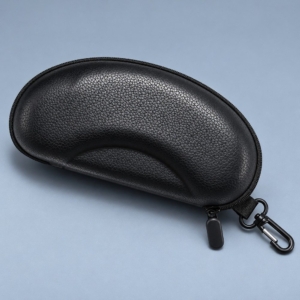 Black semi hard sunglasses case with a pebbled finish, crescent-shaped contour, zipper pull and side swivel clip on a blue-grey surface.