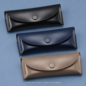 Black custom soft eyeglass case with a smooth exterior, curved flap and round fastening cap on a blue-gray background.