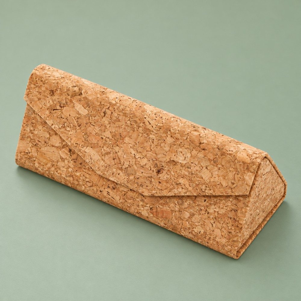 Closed tan cork-textured glasses case with an envelope-style flap and triangular profile on a sage green background.